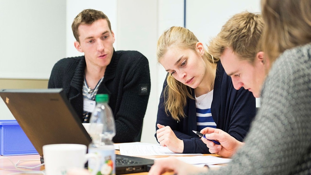 Four students collaborate around a table with laptops and notes, focused on group work in a classroom setting.