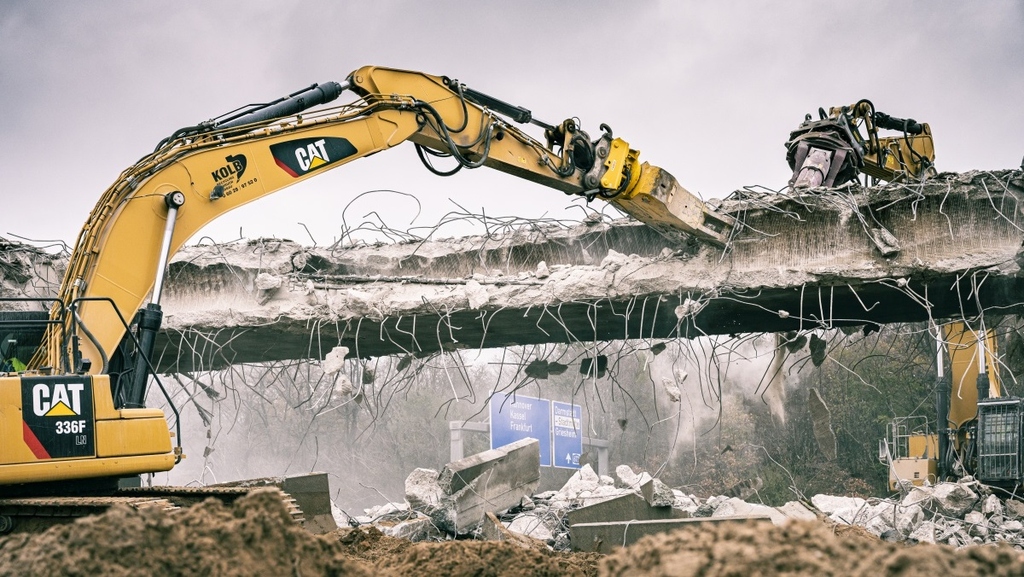 Yellow Caterpillar excavator demolishing concrete bridge with exposed rebar, dust rising, under overcast sky.